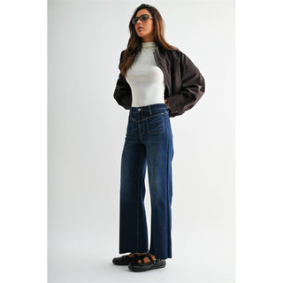A woman modeling dark blue wide-leg jeans, styled with a white ribbed turtleneck, a dark brown jacket, and black flat sandals, set against a plain light gray studio background.