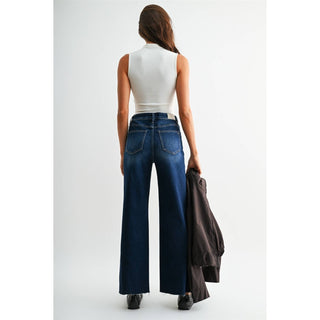 A woman is shown from behind wearing a white sleeveless top and dark blue wide-leg jeans, holding a dark brown jacket in one hand against a plain light gray studio background.