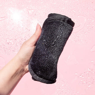 A hand holding a rolled black textured microfiber cloth against a pink background with water droplets.