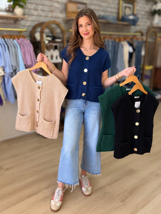 A woman in a clothing store holds up two ribbed knit cardigans on wooden hangers. The cardigan on the left is beige, and the cardigan on the right is green with a black version partially visible behind it.
