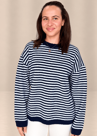 A woman wearing a navy blue and white horizontally striped knit sweater, paired with white pants, standing against a plain beige studio background.