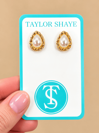 A pair of gold teardrop stud earrings with white pearl centers, displayed on a white card with a turquoise border. A hand with pink manicured nails holds the card against a solid beige background.
