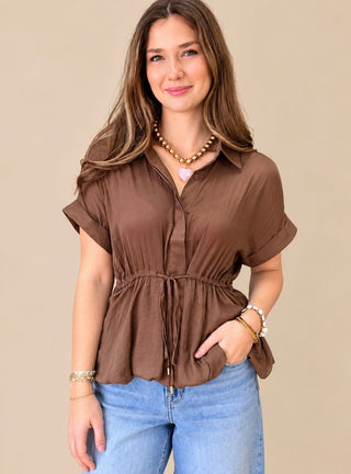 A woman wearing a brown short-sleeve button-up blouse with a gathered waist tie, paired with light blue jeans, against a solid beige background.