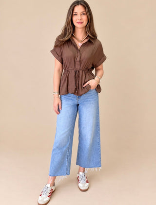 A woman modeling a brown short-sleeve button-up shirt with a drawstring waist, paired with wide-leg light blue jeans and white sneakers, against a solid beige background.