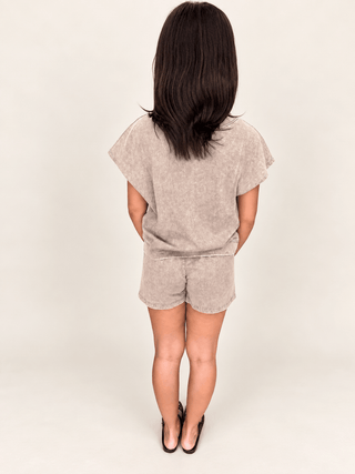 A child model shown from the back wearing a matching two-piece set consisting of a short-sleeve top and shorts in a light beige mineral-wash fabric, paired with black heels, against a plain off-white studio background.