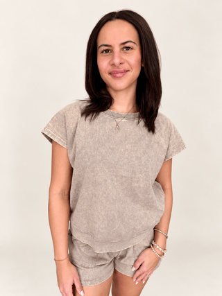 A woman modeling a matching two-piece lounge set consisting of a short-sleeve crewneck sweatshirt and shorts in a washed, mineral-washed taupe color, set against a plain light beige background.