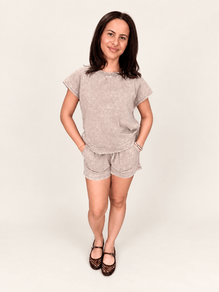A woman modeling a matching two-piece linen set consisting of a short-sleeve t-shirt and shorts in a light beige color, paired with dark patterned flats, against a plain off-white studio background.