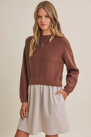A woman modeling a brown knit crewneck sweater with long sleeves, paired with a light beige pleated skirt, against a solid beige background.