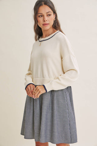 A woman modeling a cream knit sweater with dark navy trim and a navy and white vertically striped pleated skirt, set against a plain beige studio background.