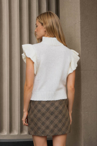 A woman shown from the back wearing a white sleeveless knit sweater vest with ruffled shoulders and a brown plaid mini skirt, standing against a stone wall.