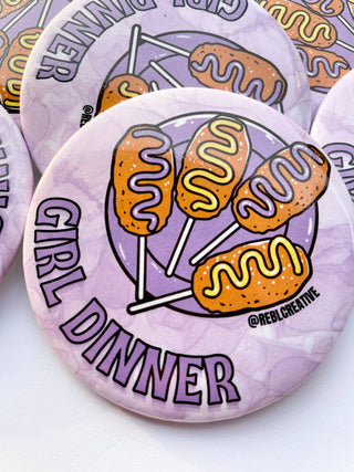 Button badge with hot dogs on sticks and 'Girl Dinner' text on a purple background