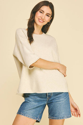 A woman wearing a cream-colored, oversized knit top with elbow-length sleeves and blue denim shorts, posing against a solid beige background.