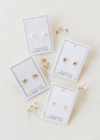 Gold earrings on LINNY CO branded cards on a beige fabric background
