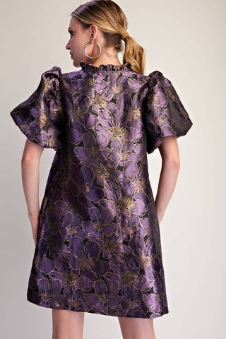 A woman shown from the back wearing a short-sleeved purple dress with a metallic gold floral jacquard pattern, photographed against a neutral light beige background.