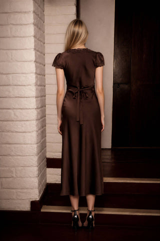 A woman viewed from behind wearing a dark brown midi dress with short flutter sleeves, lace detailing at the neckline, and a tie waist, standing on wooden stairs next to a white brick column.