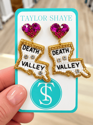 A hand holding a turquoise card displaying a pair of statement earrings shaped like the state of Louisiana, with 'DEATH VALLEY' text, gold-tone outlines, white textured centers, and bright pink heart-shaped rhinestones. The card reads 'TAYLOR SHAYE' at the top.