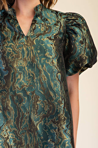 A close-up of a model wearing a short-sleeve blouse with a teal, black, and gold abstract marbled pattern.
