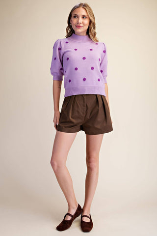 A woman modeling a lavender knit mock-neck sweater with purple polka dots and brown pleated shorts against a solid beige studio background.