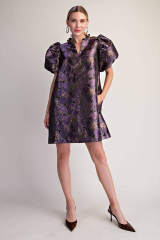 A woman modeling a short, knee-length purple dress with a metallic gold floral jacquard pattern, puffed short sleeves, and a high collar, paired with dark brown pointed-toe heels against a light gray studio background.