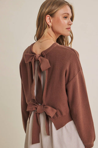 A woman wearing a brown knit sweater with a large bow detail on the back, paired with a cream and brown striped dress, photographed against a plain beige background.