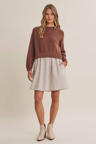A woman modeling a two-piece outfit consisting of a brown long-sleeve crewneck sweater and a light beige pleated mini skirt, paired with beige ankle boots, against a solid light beige studio background.