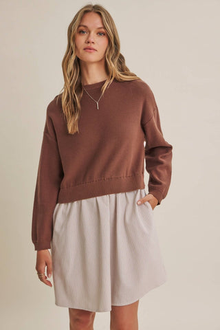A woman wearing a brown knit crewneck sweater and a light beige pleated skirt, standing against a plain beige studio background.