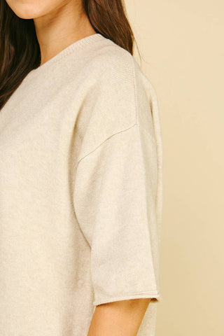 Close-up of a person wearing a light cream knit short-sleeve top against a warm beige background.