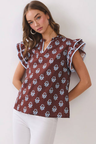 A woman modeling a short-sleeve blouse with flutter sleeves, featuring a brown base with white floral block prints and thin vertical stripes, paired with white pants against a plain white studio background.