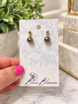 A pair of gold drop earrings with dark gray pearl-like beads, displayed on a white floral backing card held by a hand with pink nail polish. The card is branded 'Blue Blossom'.