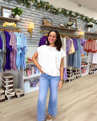 A woman stands in a clothing boutique wearing a white short-sleeve top and light blue wide-leg jeans. The store has a gray wood-paneled wall, floral garlands, racks of colorful clothing, and shelves with shoe boxes.