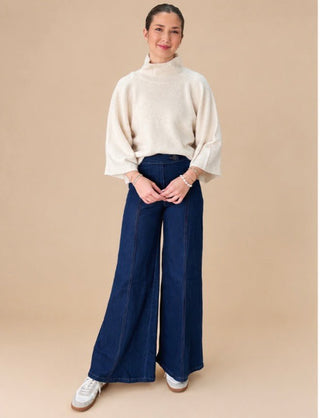A woman modeling a cream-colored knit mock-neck sweater with wide bell sleeves and dark blue wide-leg denim pants, standing against a solid beige studio background.
