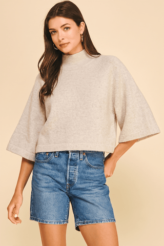 A woman modeling a light beige knit mock-neck poncho sweater with wide flared sleeves, paired with blue denim shorts, against a solid beige background.