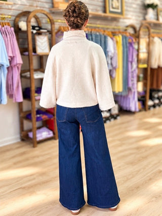 A woman viewed from behind wearing a cream-colored fuzzy mock-neck sweater and dark blue wide-leg denim jeans, standing in a clothing boutique with racks of colorful garments in the background.