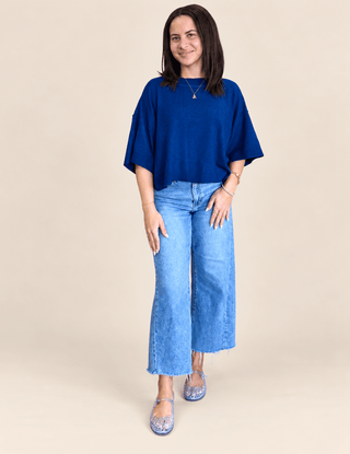 A woman modeling a loose-fitting royal blue short-sleeve top paired with light-wash blue wide-leg jeans and purple patterned flats, set against a solid beige studio background.