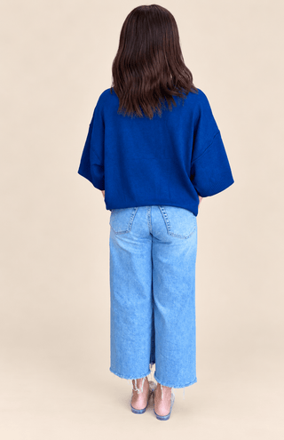A woman viewed from behind wearing a vibrant royal blue oversized short-sleeve top, light-wash wide-leg denim jeans, and pink high heels, set against a solid beige studio background.