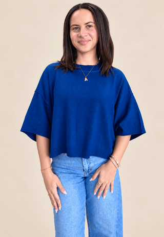 A woman wearing a vibrant royal blue knit sweater with wide, elbow-length sleeves and light wash blue jeans, standing against a solid beige background.