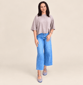 A woman modeling a light gray short-sleeve crop top and wide-leg blue jeans against a solid beige studio background.