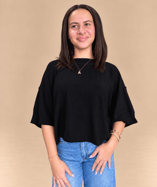A woman wearing a black short-sleeve knit top and blue jeans, standing against a plain beige studio background.