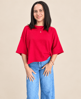 A woman modeling a bright red knit sweater with wide three-quarter sleeves and blue jeans, standing against a solid beige background.