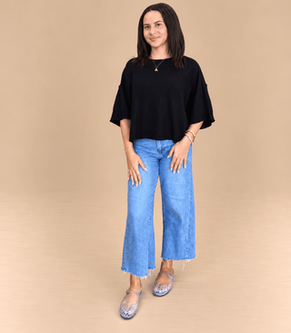 A woman modeling a black short-sleeve top and light blue wide-leg jeans against a solid beige studio background.