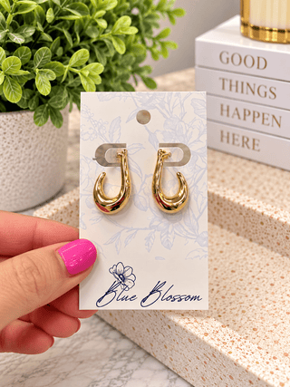 A hand with bright pink nail polish holds a pair of small gold hoop earrings on a white display card. The card is branded 'Blue Blossom' and is photographed in a styled indoor setting with a potted plant and stacked books in the background.