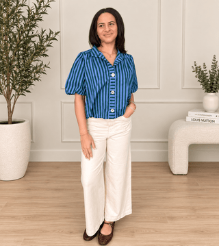 A woman stands in a bright indoor room wearing a blue and navy vertically striped short-sleeve button-up shirt with cream wide-leg pants and dark brown flats.