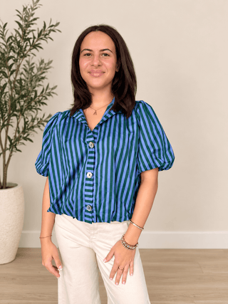 A woman modeling a blue and green vertically striped short-sleeve blouse with puff sleeves, paired with cream pants, standing in a brightly lit room with a potted plant and light wood floor.