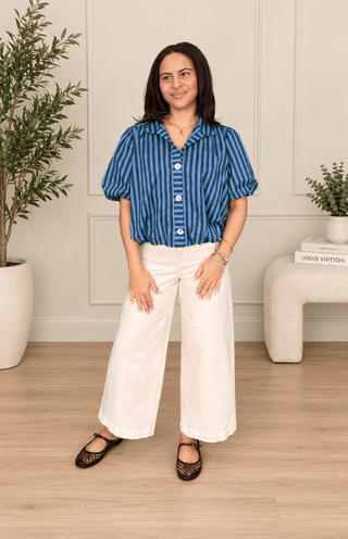 A woman modeling a blue and black vertically striped short-sleeve button-up shirt with white wide-leg pants and black lace-up flats, standing in a bright room with a light wood floor, white wall paneling, and potted plants.