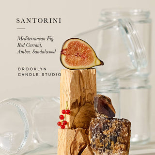 A candle product graphic with the text 'SANTORINI' and 'Mediterranean Fig, Red Currant, Amber, Sandalwood' above a styled arrangement of wood pieces, a sliced fig, red berries, and blurred glass jars on a warm beige background.