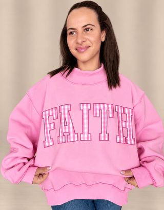 A woman wearing a pink mock-neck sweatshirt with the word 'FAITH' in pink-and-white gingham block letters outlined in black, posed against a neutral beige background.
