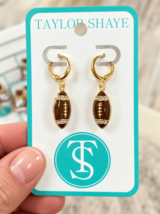 A pair of gold hoop earrings with football-shaped charms, held on a white display card with turquoise trim. A hand is visible holding the card, and the card is branded Taylor Shaye with a large TS monogram.