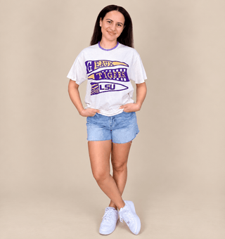 A woman modeling a white graphic t-shirt with purple and gold LSU 'GAWK TIGHS' lettering, paired with light blue denim shorts and white sneakers, against a solid beige studio background.