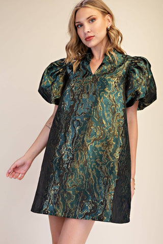 A woman modeling a short puff-sleeve mini dress with a green, gold, and black marbled jacquard pattern against a solid beige studio background.