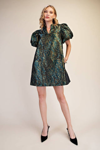 A woman modeling a short puff-sleeve mini dress with an abstract marbled pattern in dark teal, green, and gold, paired with black pointed-toe heels against a plain beige studio background.